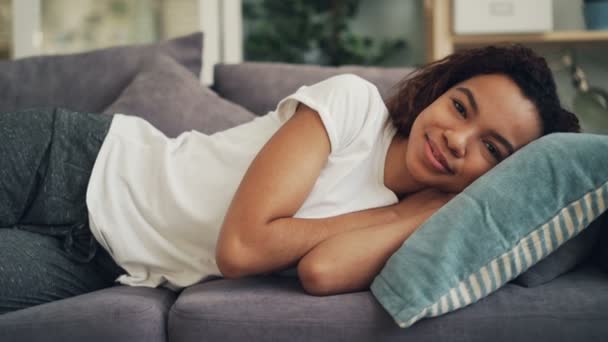 Retrato de una dama afroamericana sonriente tumbada en un sofá en casa con la cabeza sobre la almohada y mirando a la cámara. Descanso diurno, jóvenes y concepto de estilo de vida . — Vídeo de stock