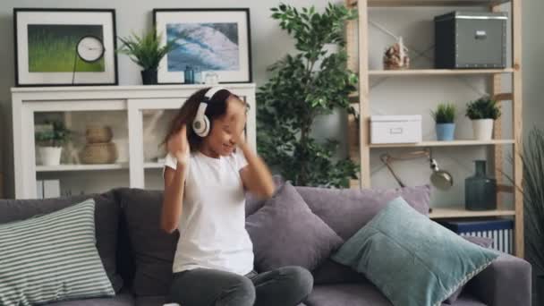 Happy African aAmerican dama en ropa casual es escuchar música con auriculares, bailar y relajarse sentado en el sofá en casa. Tecnología moderna y concepto juvenil . — Vídeo de stock