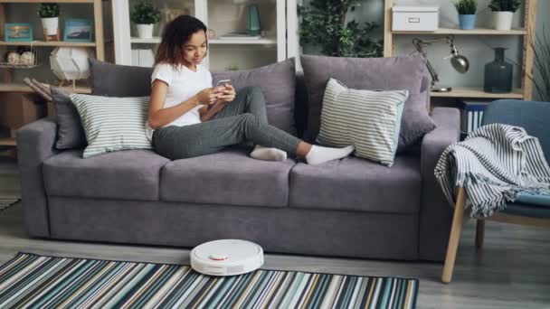 Jonge African American vrouw is met behulp van slimme telefoon en ontspannen op de bank terwijl robot stofzuiger is stofzuigen appartement. Moderne technologieën en huishoudelijke concept. — Stockvideo