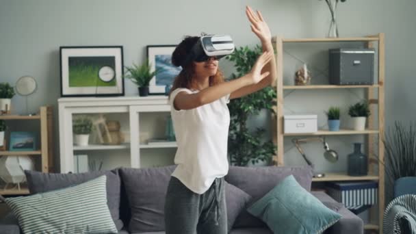 Young African American woman is enjoying experience with augmented reality glasses wearing modern vr headset. Girl is moving hands and head standing at home. — Stock Video