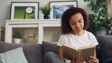 Yakışıklı Afro-Amerikan öğrenci kitap okuma ve gülümseyen sonra rahat kanepe yalnız güzel ışık dairede oturan sayfa dönüm. Gençlik ve eğitim kavramı.