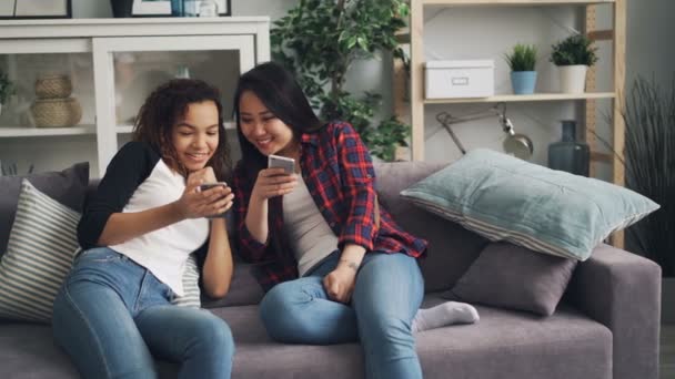 Des étudiants joyeux asiatiques et afro-américains parlent et rient en regardant l'écran du smartphone à l'aide de gadgets assis sur le canapé à la maison. Concept jeunesse et dispositifs . — Video