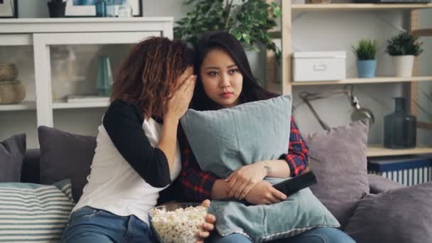 Emotional young women are watching horror film together hiding behind pillows and closing eyes. Girls are eating popcorn sitting on sofa in modern apartment. — Stock Video