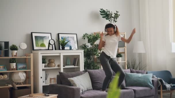 Un adolescent afro-américain actif danse sur un canapé en écoutant de la musique à travers un casque et en s'amusant seul à la maison. Loisirs, jeunesse et chansons concept . — Video