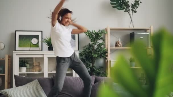 Lekfulla afroamerikanska kvinnan hoppar dans på soffan på hem lyssnar på musik med hörlurar som njuter av rytm och melodi. Ungdomskoncept livsstil och kultur. — Stockvideo