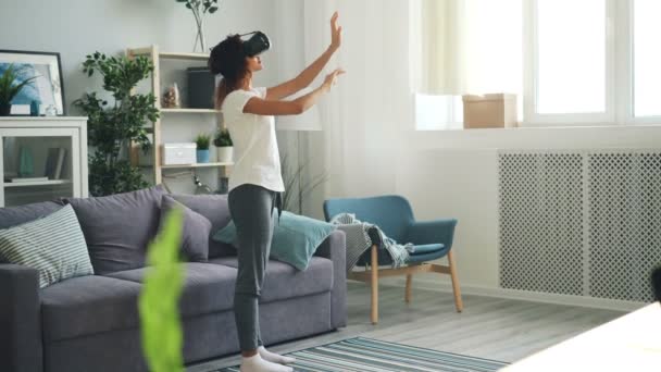 Mujer joven delgada en ropa casual está utilizando gafas de realidad aumentada de pie en la sala de luz en casa y mover las manos tocando el aire. Dispositivos y concepto de entretenimiento . — Vídeo de stock