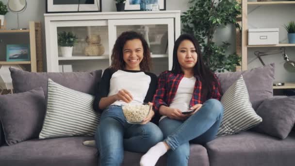 Happy female friends are watching funny movie together laughing, talking and eating popcorn sitting on couch at home. Friendship and television concept. — Stock Video