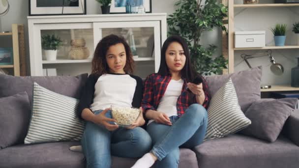 Bored young women are watching TV together at home and eating popcorn sitting on sofa in living room. Asian girl is holding remote control and pushing buttons. — Stock Video