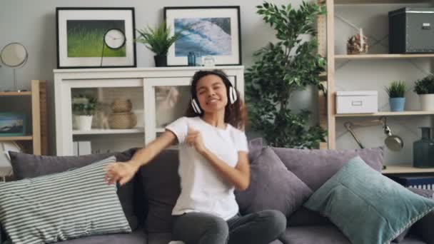 Cute African American girl is listening to music using headphones, singing and dancing sitting on couch in modern apartment. Beautiful people and hobby concept. — Stock Video