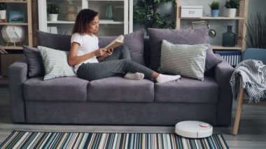 Rahat Afro-Amerikan kız robot elektrik süpürgesi zemin içinde yerine kişinin ev süpürmek iken koltukta istirahat kitap okuyor. Gadget'lar ve ev kavramı.