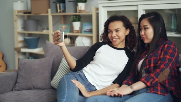 Mujeres jóvenes sonrientes Asiático y afroamericano haciendo videollamada en línea mirando la pantalla del teléfono inteligente hablando y riendo sentado en el sofá en un bonito apartamento . — Vídeo de stock