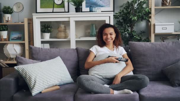 Zoom-in docela africký americký student sledování legrační komedie v televizi a směje se sedí na pohovce a držení dálkového ovládání. Koncept televize a emoce. — Stock video