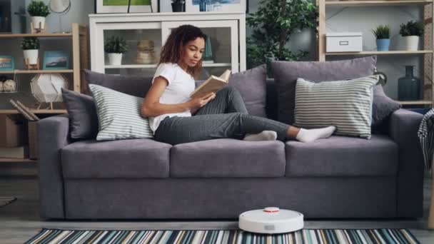 Snygga afrikanska amerikansk student vänder på robotiserade dammsugare och läsebok njuter av hobby och kopplar av på soffan medan gadget städa golvet. — Stockvideo