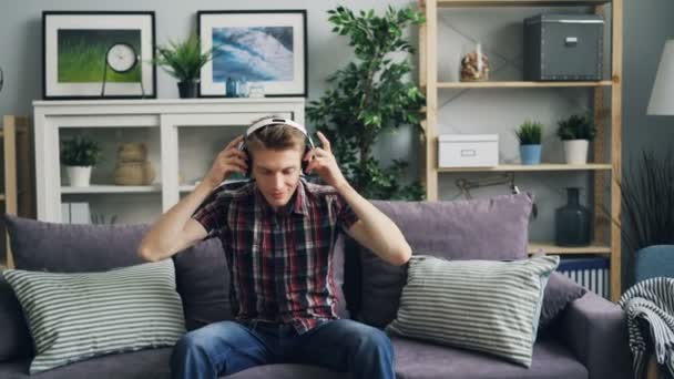 Збільшення щасливого хлопця меломанка одягає навушники і слухає музику і посміхається насолоджуючись мелодією і ритмом, сидячи на дивані. Молодіжна культура та концепція способу життя . — стокове відео