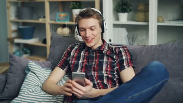 陽気な学生は、スマート フォンの画面と笑って楽しんで、ヘッドホンで音楽を聴くことを見て使用しています。デバイスおよび青年の近代的な概念. — ストック動画