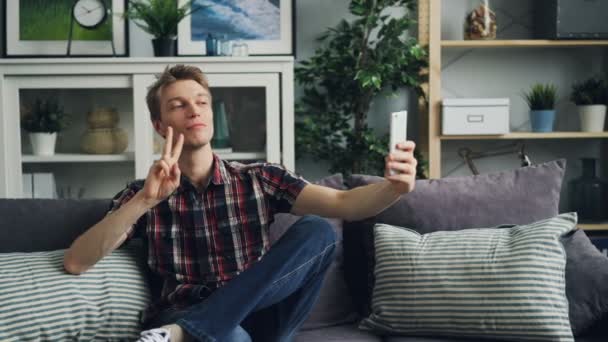 Jovem bonito está tirando selfie com câmera de telefone inteligente posando com gestos de mão na moda dedais-up, arma, OK e v-sign sentado no sofá em bom apartamento . — Vídeo de Stock