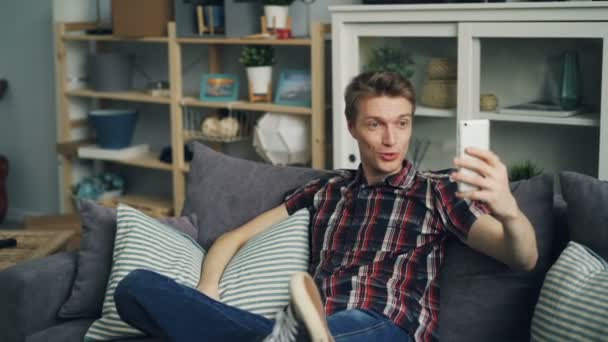 즐거운 젊은 남자 집에서 소파에 앉아 화면을 보고 웃 고 얘기 하는 스마트폰을 사용 하 여 온라인 영상 통화를 하 고 있습니다. 사람들 및 통신 개념. — 비디오