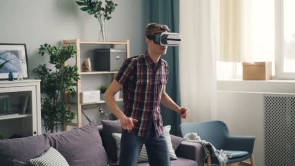 Adult man in casual clothing is concentrated on game in virtual reality glasses standing at home wearing device and moving hands. AR experience and youth concept. — Stock Video