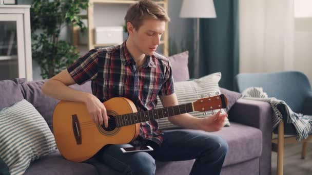 Νεαρός μουσικός tuning ακουστική κιθάρα αγγίζει χορδές που κάθεται στον καναπέ στο σπίτι κατά τη διάρκεια του ελεύθερου χρόνου. Νεολαία lifestyle, διαμερίσματα και μουσική έννοια. — Αρχείο Βίντεο