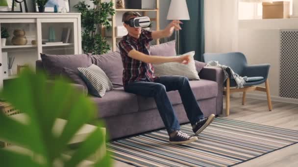 Sorglosigkeit Student spielt Spiel mit Virtual-Reality-Brille fahren Autorennen sitzt auf dem Sofa zu Hause und bewegt Hände und Beine. Freizeit- und Spaßkonzept. — Stockvideo