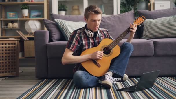 Jovem em roupas casuais está assistindo tutorial usando laptop e tocando guitarra sentado no chão em casa aprendendo a usar instrumento musical. Conceito de juventude e internet . — Vídeo de Stock