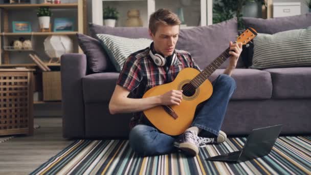 Giovane uomo in abbigliamento casual sta guardando tutorial utilizzando il computer portatile e suonare la chitarra seduto sul pavimento a casa imparare a utilizzare lo strumento musicale. Concetto di gioventù e internet . — Video Stock