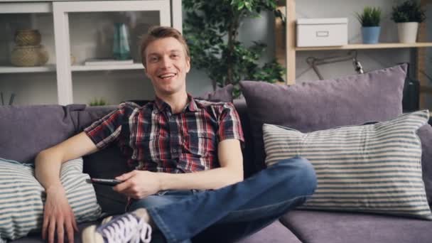 Vrolijke mannelijke student is grappige sitcom op Tv kijken en lachen, ontspannen op de Bank en houden van de afstandsbediening. Televisie en positieve emoties concept. — Stockvideo