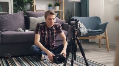 Duygusal genç adam blogger kulaklık giyen ve tripod üzerinde kamera ile katta oturan sanal gerçeklik gözlük hakkında video kaydediyor. Bloglama ve Gadget'ları kavramı.