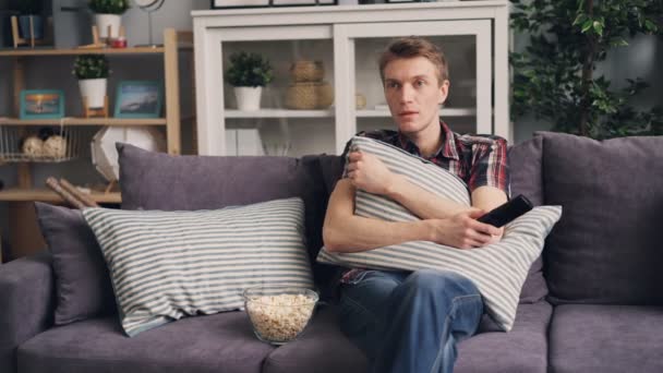 感情的な若い男は、ポップコーンを食べたり、リモート コントロールを保持しているソファに座っている枕の後ろに隠れて、自宅テレビでホラー映画を見ています。エンターテイメント、テレビのコンセプト. — ストック動画