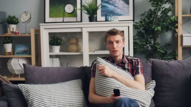 Zoom-in emotionaler junger Mann weint über traurigen Film im Fernsehen sitzt auf bequemem Sofa und hält Fernbedienung und Kissen in der Freizeit zu Hause. — Stockvideo