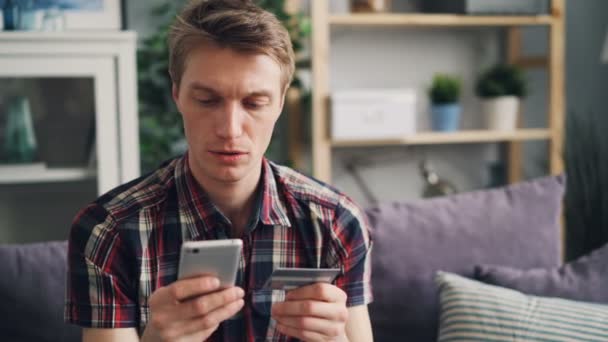 Estudiante guapo está de compras en línea haciendo el pago con tarjeta de crédito con pantalla táctil smartphone y luego sonriendo en casa. Finanzas, comercio electrónico y concepto de juventud . — Vídeo de stock