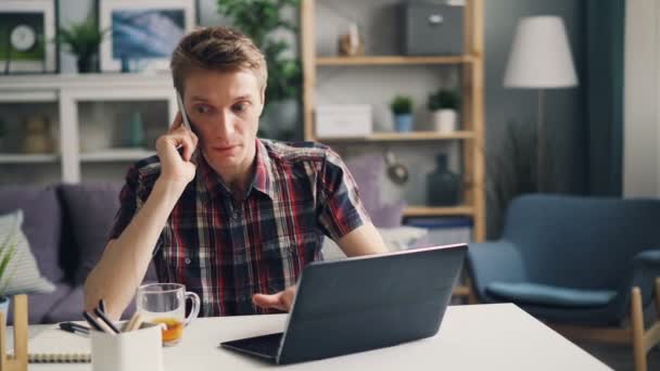 Geïrriteerde man freelance medewerker is praten over mobiele telefoon en laptop uiten van negatieve emoties werken in appartement alleen gebruikt. Contacten en gevoelens bedrijfsconcept. — Stockvideo