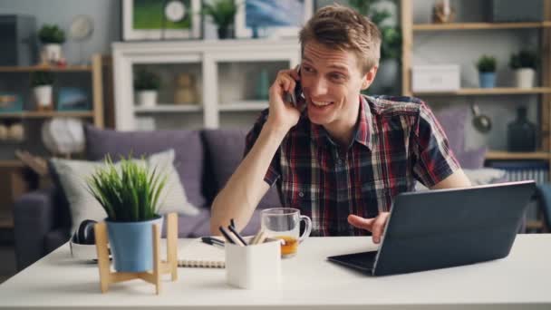 Χαρούμενος νεαρός freelancer μιλάει στο κινητό και πληκτρολόγησης λειτουργεί σε απευθείας σύνδεση χρησιμοποιώντας το laptop κάθεται στο χώρο εργασίας στο σπίτι. Χιλιετία και επιχειρηματική αντίληψη. — Αρχείο Βίντεο