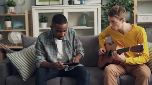 Kaukasische jonge man en zijn Afro-Amerikaanse vriend leren om te spelen de gitaar thuis met behulp van Tablet PC tijd samen doorbrengen. Hobby, muziek en technologie concept. — Stockvideo