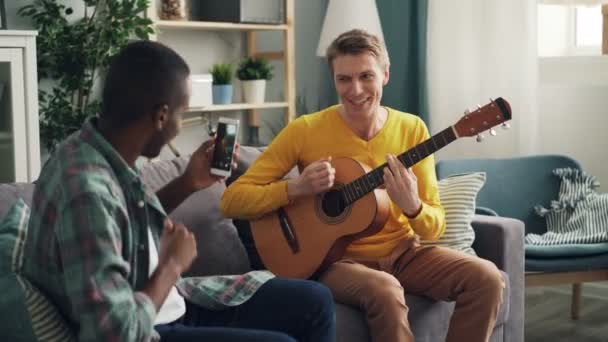 El joven afroamericano sostiene un teléfono inteligente moderno y hace un video de su amigo caucásico tocando la guitarra y cantando en casa. concepto de diversión y gadgets . — Vídeos de Stock