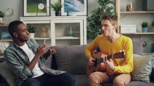 Los amigos masculinos se están divirtiendo en casa, el chico caucásico está tocando la guitarra y el joven afroamericano está grabando video con teléfono inteligente. Concepto de amistad y música . — Vídeo de stock