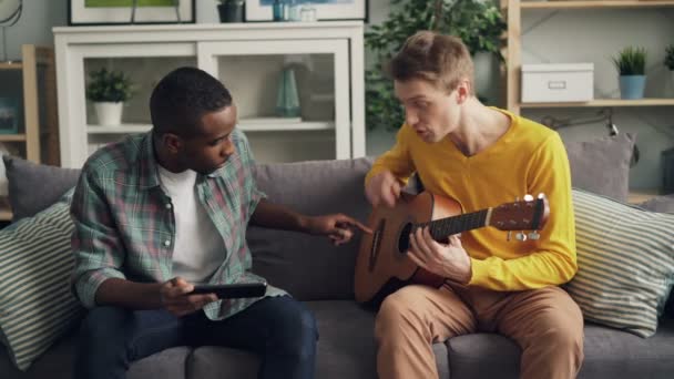 Chico afroamericano está usando tableta para enseñar a su amigo caucásico a tocar la guitarra aprendiendo en casa juntos. Los hombres están hablando y tocando instrumento musical disfrutando de la actividad . — Vídeos de Stock