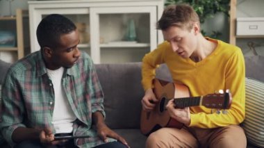 Mutlu genç erkekler beyaz ve Afro-Amerikan olan evde eğlenceli gitar çalmak ve kullanma zevk müzik ve ritim tablet. Gençlik ve hobi kavramı.
