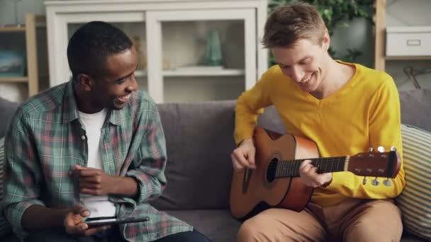 Vrolijke jongens Afrikaanse-Amerikaanse en Kaukasische vrienden zijn gitaar te spelen, met behulp van tablet en lachen ontspannen op de Bank samen in modern appartement. Vriendschap en muziek concept. — Stockvideo