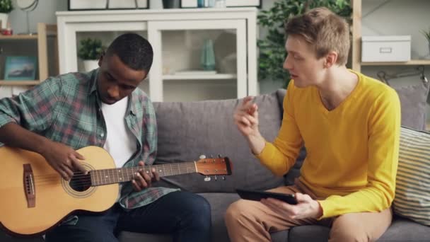 Bonito cara está ensinando seu amigo afro-americano para tocar guitarra usando tablet, os homens estão sentados no sofá olhando para a tela e tocando cordas no instrumento musical . — Vídeo de Stock