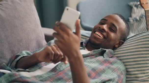 Bonito afro-americano cara está se divertindo com smartphone tocando tela deitada no sofá em casa passar tempo de lazer com o dispositivo. Millennials e conceito de tecnologia . — Vídeo de Stock