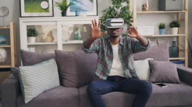 Afrika kökenli Amerikalı adam gülümseyerek ve kanepede oturan ve yeni bir deneyim zevk eller hareket gülüyor sanal gerçeklik gözlük kullanıyor. Millennials ve aygıtları kavramı.
