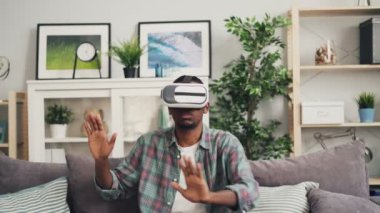 Zoom-in Afrika kökenli Amerikalı gencin giyen augmented gerçeklik gözlük ve hareketli el gülümseyen ve emjoying oyun kanepe modern dairede oturan.