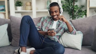 Yakışıklı bir Afrikalı-Amerikalı adam rahat giyim kulaklık müzik dinlemek ve şarkı evde kanepede oturan ve smartphone kullanarak. Gadget'lar ve gençlik kavramı.