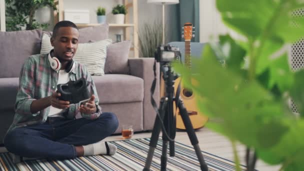 Blogger joven afroamericano está grabando video sobre gafas de realidad aumentada sentadas en el suelo hablando con auriculares y usando cámara en el trípode . — Vídeo de stock