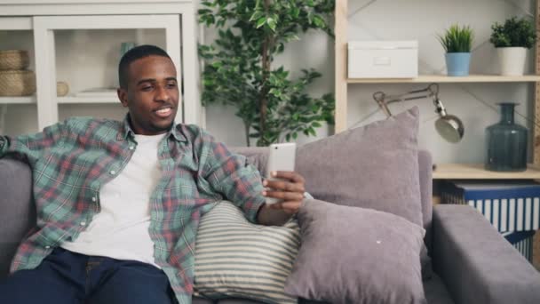 笑顔のアフリカ系アメリカ人の男は、オンライン ビデオ通話の話と、ソファに座ってスクリーンを見て笑っているようにスマート フォンを使用しています。人とコミュニケーションの概念. — ストック動画