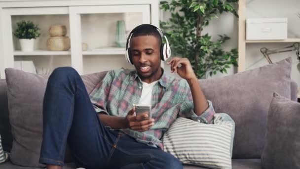 Alegre chico afroamericano está cantando y escuchando música en los auriculares relajarse en el sofá en el apartamento moderno disfrutando de la canción. Cultura juvenil y concepto de estilo de vida . — Vídeo de stock