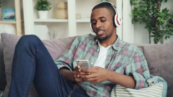 Estudante afro-americano atraente está usando a tela de toque do smartphone e ouvir música com fones de ouvido no sofá em casa. gadgets modernos e conceito millennials . — Vídeo de Stock