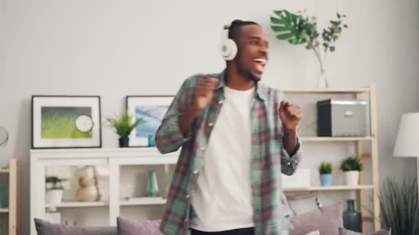 Fröhliche afrikanisch-amerikanische Studentin tanzt singend und hört Musik über kabellose Kopfhörer, die es sich zu Hause gemütlich machen. Jugendkultur und Lifestyle-Konzept. — Stockvideo