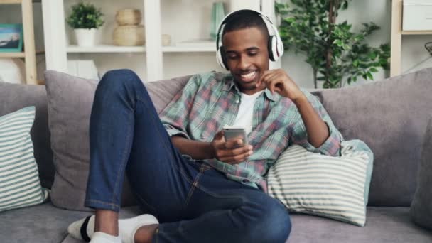 Un bell'uomo afroamericano in abbigliamento casual sta ascoltando musica in cuffia e cantando seduto sul divano a casa e usando lo smartphone. Gadget e concetto di gioventù . — Video Stock